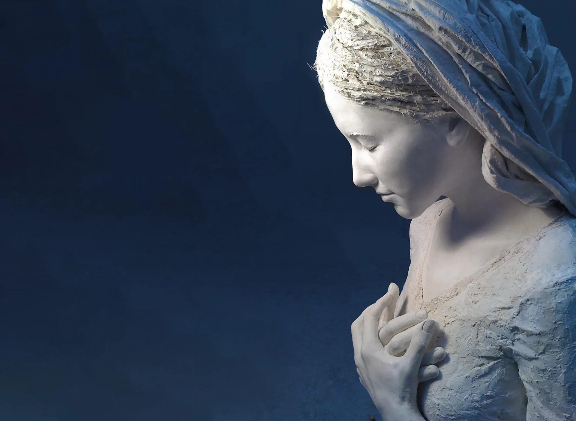 Escultura de la Virgen en oración, con velo, sobre fondo azul noche